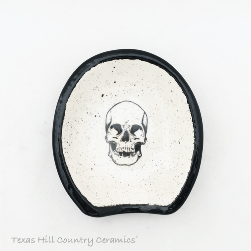 Oval shape ceramic spoon rest with skull face and black trim.